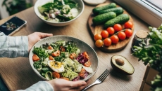 Hands hold a bowl of salad, with fresh vegetables laid out on a table