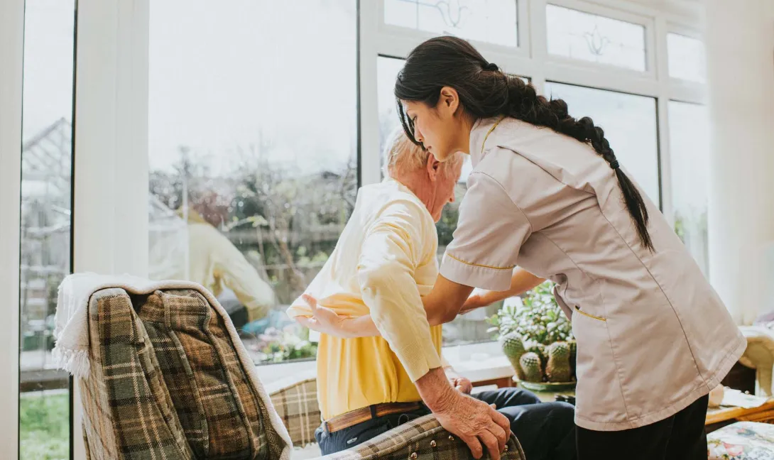 A nurse helps a resident into a chair. A nurse helps a resident into a chair.
