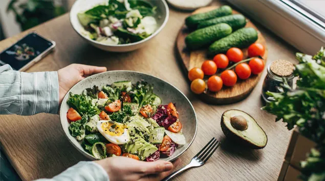 Hands hold a bowl of salad, with fresh vegetables laid out on a table